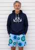 Man wearing a navy hoodie with anchor design and blue floral shorts against a white wall.