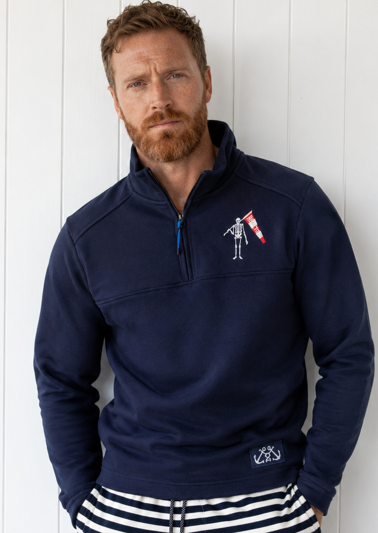 Man wearing a navy blue pullover with a logo and striped shorts against a white wooden background