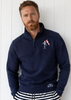 Man wearing a navy blue pullover with a logo and striped shorts against a white wooden background