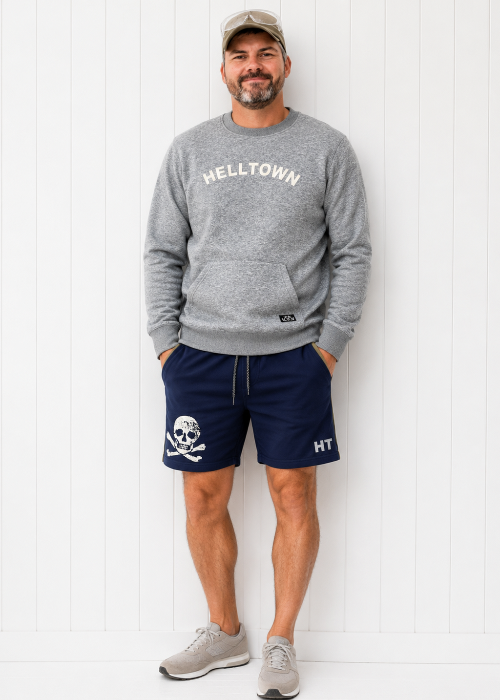 Man wearing a gray 'HELLTOWN' sweatshirt and navy shorts with a skull design, standing against a white wooden panel background.