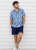 A man wearing a light blue short-sleeve button-up shirt with a colorful abstract print, navy blue shorts, and white sneakers, standing against a white horizontal siding wall.