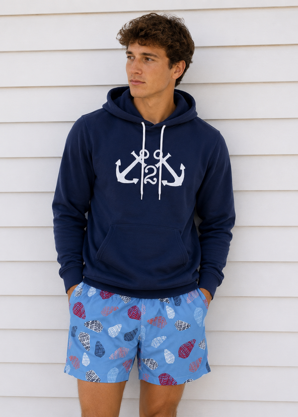 Man wearing a navy hoodie with anchor design and colorful shorts against a white wall.