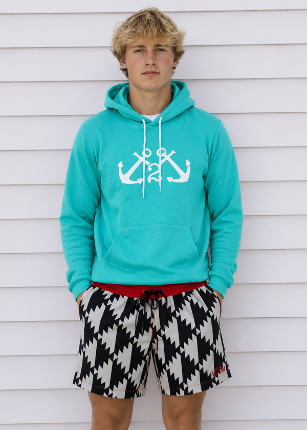 Person wearing a turquoise hoodie with anchor design and black and white patterned shorts against a white wooden background