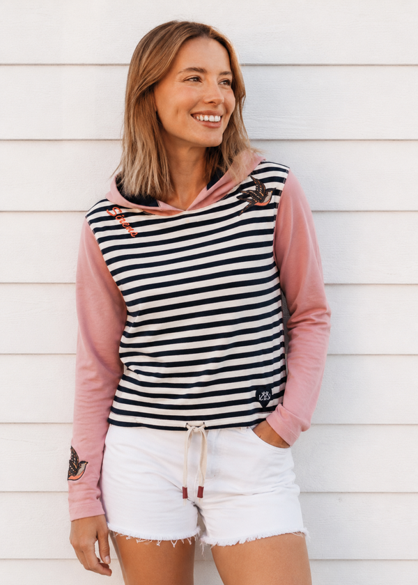 A woman wearing a pink and black-and-white striped hooded top with white shorts, standing against a white wood-paneled wall.