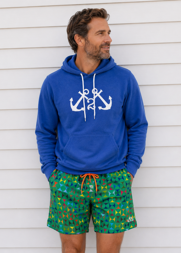 Man wearing a blue hoodie with anchor design and colorful shorts against a white wall.