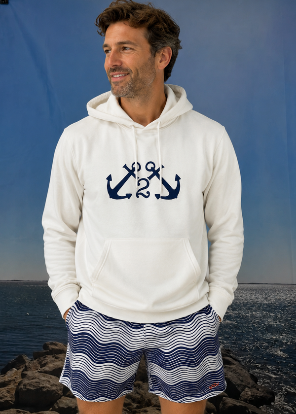Man wearing a white hoodie with an anchor design and striped shorts standing by a body of water.